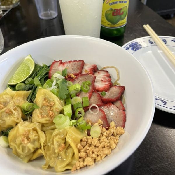 Thai dish with sliced red pork, wontons, green onions, and lime, with a drink and chopsticks.