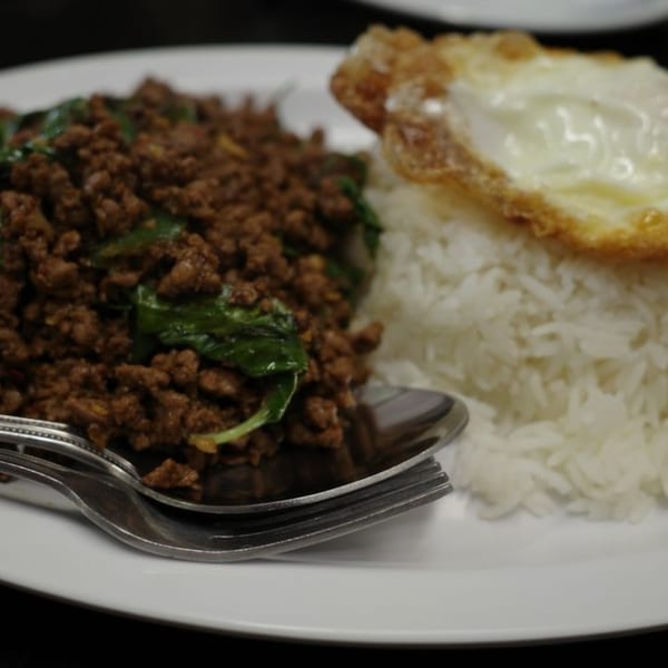 Thai basil pork with rice and a fried egg on a white plate with silverware.