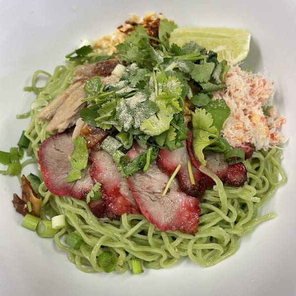 Green noodles topped with sliced pork, duck, crab meat, cilantro, and a lime wedge.